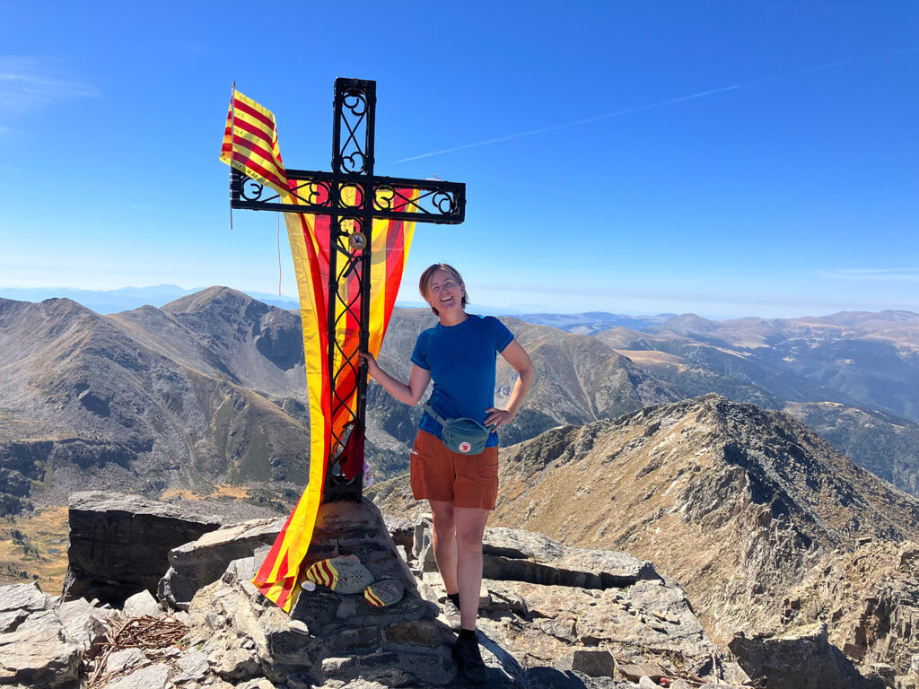 Me at the top of Pic du Carnigou, holding a cross with two yellow and red flags and with beutiful mountains in the back
